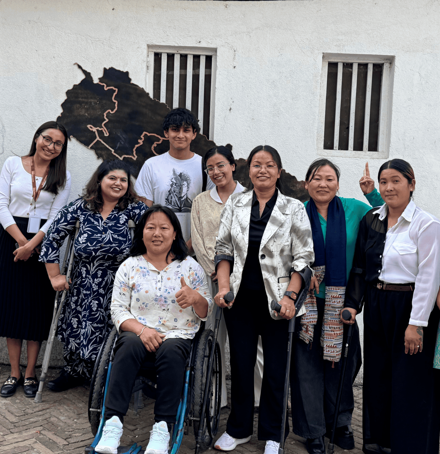 A diverse group of eight people, including two women in wheelchairs, pose together and smile in front of a white wall with a metal map art installation. The group includes both men and women, dressed in a mix of casual and professional attire, suggesting a friendly or inclusive event setting.